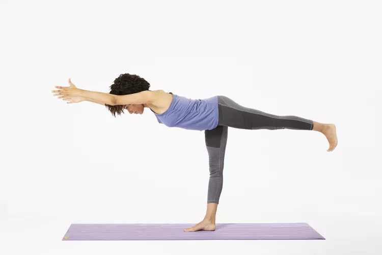 Une femme réalise la posture du guerrier III sur un tapis de yoga.