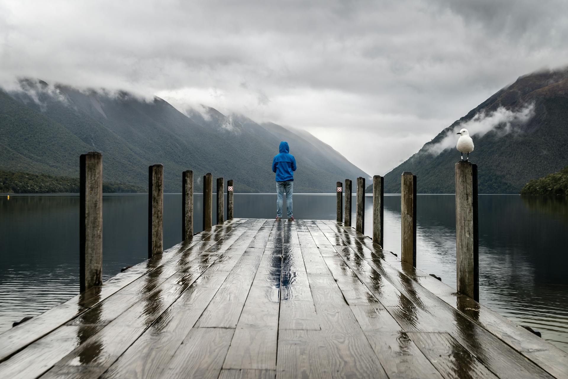Un homme avec un manteau imperméable sous la pluie, au bout d'un ponton face à un lac et aux montagnes.
