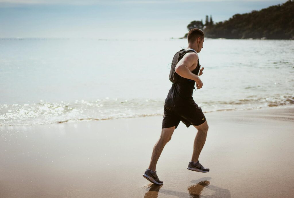 Un homme pratique le trail running sur une plage près de l'eau, en portant un sac d'hydratation.